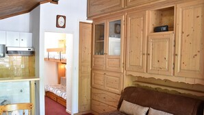 Een knus ingerichte kamer met een houten bankje, een bruine bank en een keukenhoek met kastjes en een klok.