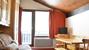 Een gezellige kamer met een houten plafond, een bank, een tafel met stoelen, een televisie en een raam met gordijnen.