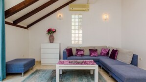 Un salon avec un canapé bleu, une table basse blanche, un banc bleu, un meuble blanc, une fenêtre à motifs géométriques et un appareil de climatisation.