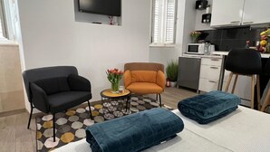 A modern living room with a black sofa, an orange armchair, and a small round table with a vase of flowers. There is a flat-screen TV mounted on the wall, a kitchen area with cabinets and a microwave, and a bed with blue bedding. - Apartments Noa Old Town - Studio - First Floor (Dubrovnik)