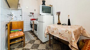 Uma cozinha compacta com geladeira, fogão e uma pequena televisão em cima. Há uma cadeira de madeira com almofada colorida e uma mesa com toalha estampada.