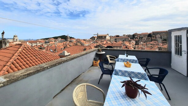 Terraço no telhado com mesa e cadeiras, um vaso de planta e vista para paisagem urbana com telhados de terracota e edifícios históricos.