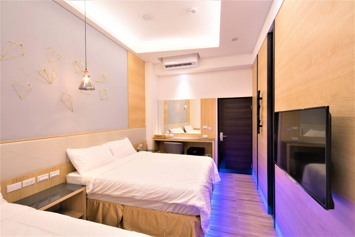 A hotel room with two beds, a bedside table, a mirror, a TV, and a ceiling light.