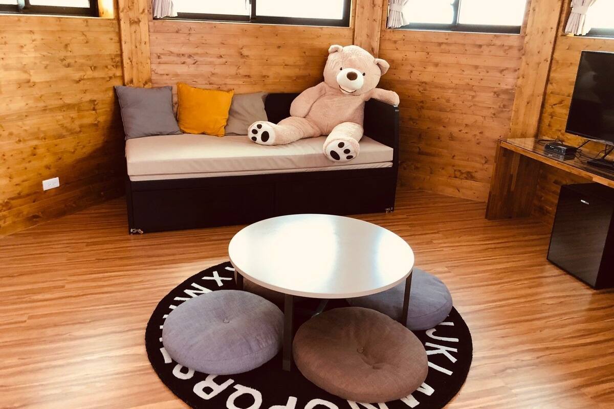 A cozy room with a wooden interior, a sofa with cushions, a large teddy bear, a small round table, and a television on a wooden stand.