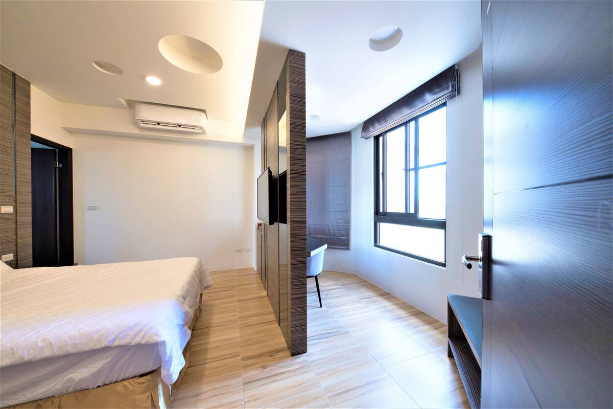 A modern bedroom with a bed, a built-in wardrobe, a window with blinds, and an air conditioning unit.
