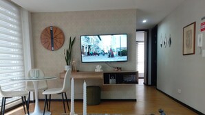 Ett modernt vardagsrum med ett soffbord i glas, vita stolar, ett sidobord i trä med dekorativa föremål, en platt-TV som är monterad på väggen och en stor väggklocka.