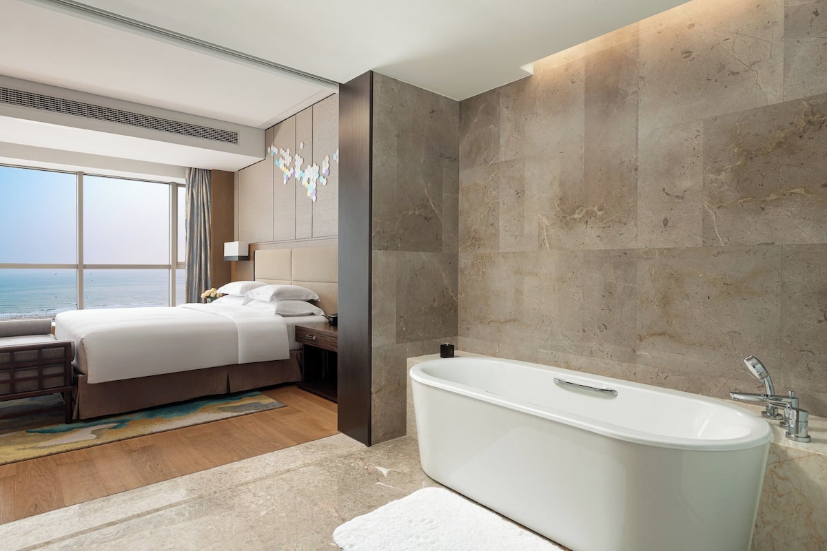 A modern hotel room with a large bed, a bathtub, and a view of the sea.