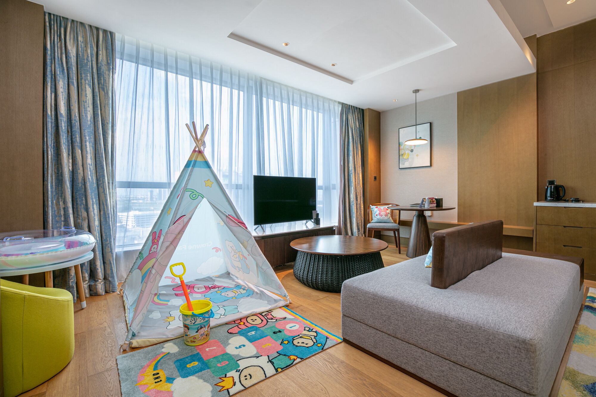 A modern hotel room with a cozy living area, a flat-screen TV, a dining table, and a colorful play mat.