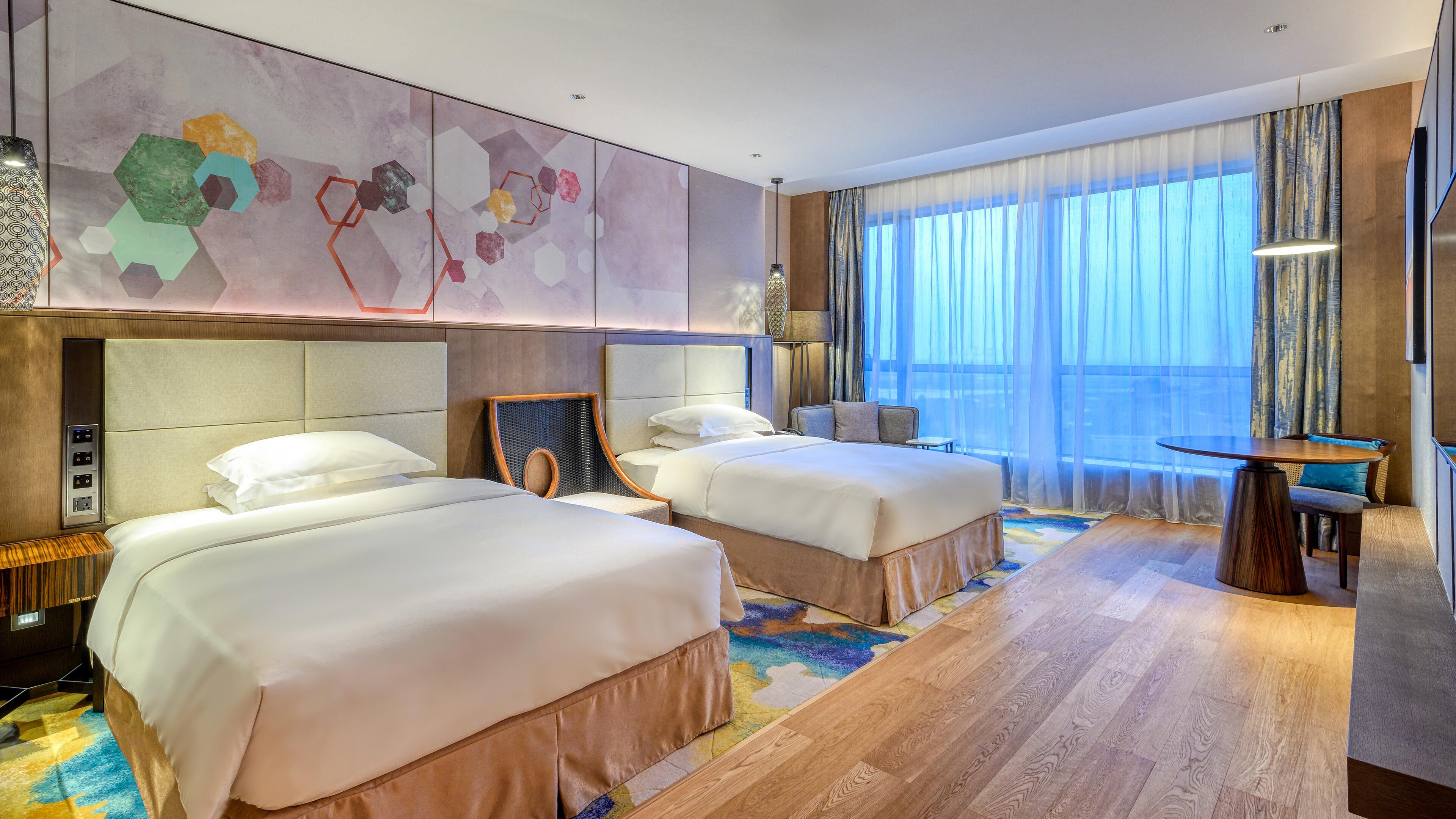 A hotel room with two beds, a large window with sheer curtains, a wooden floor, and a wall with abstract art.