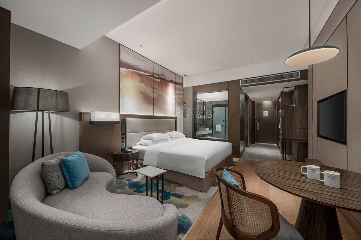 A modern hotel room with a bed, a seating area with a table and chairs, a desk, a lamp, and a TV.