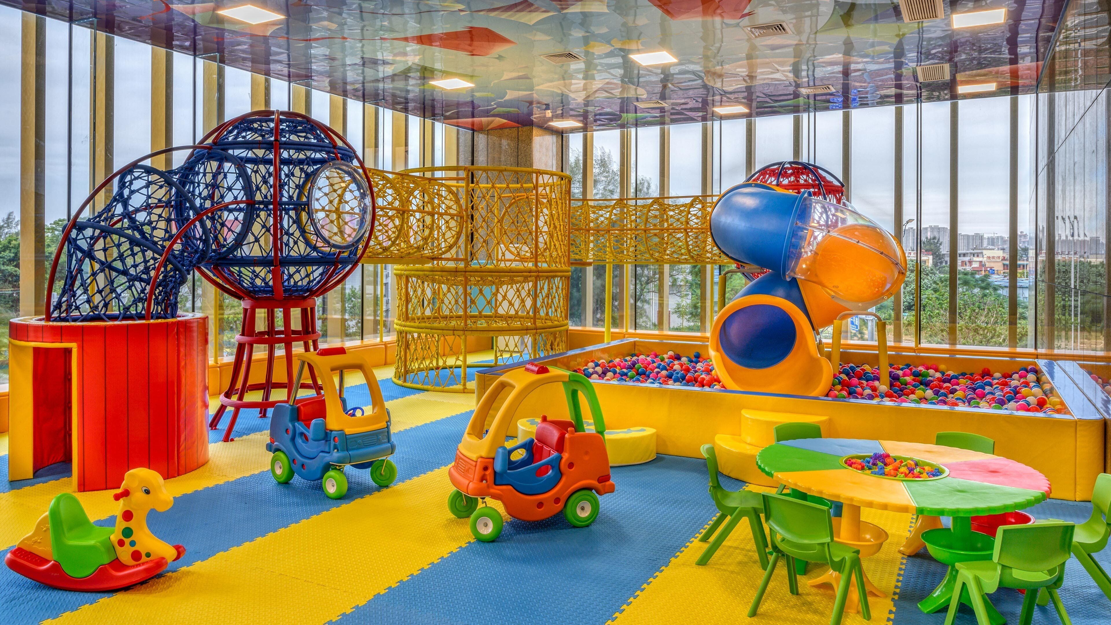 Ein Kinderspielbereich mit verschiedenen Spielgeräten, einer Bällecke und Blick auf die Stadt durch große Fenster.