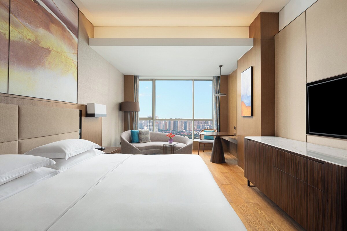 Una habitación de hotel moderna con una cama grande, un sofá, una mesita, un televisor y vistas a la ciudad.