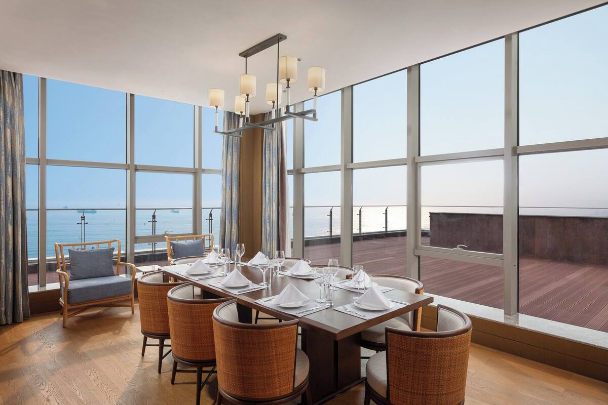 A dining area with a large table set for six, large windows, and a view of the sea.
