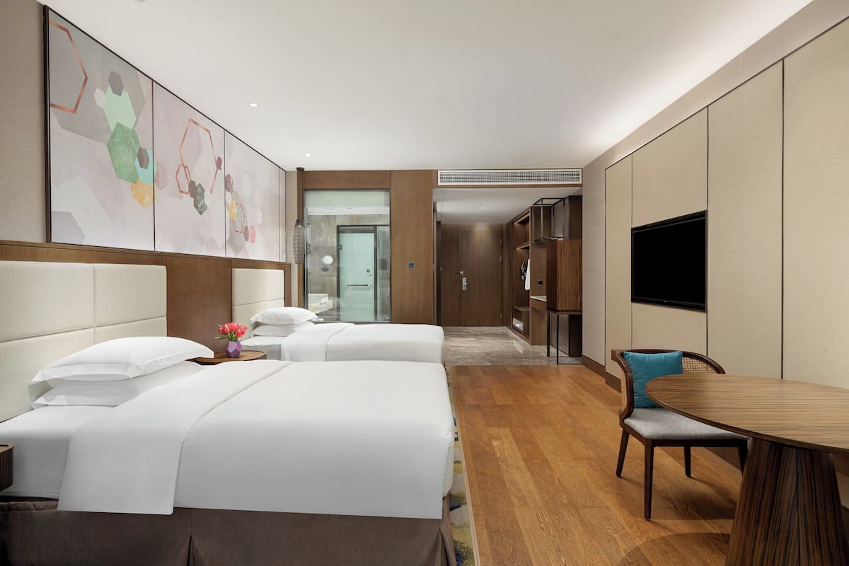 A hotel room with two beds, a wooden dining table, a TV, and a bathroom visible in the background.
