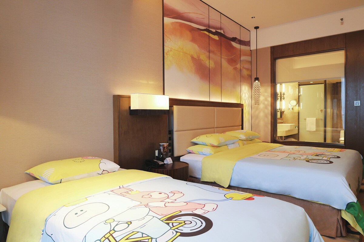 Habitación de hotel con dos camas, cada una con diseños de personajes de dibujos animados en la ropa de cama. Hay una mesita de noche con una lámpara, un espejo y una pequeña mesa con un teléfono y una revista. La habitación tiene un ventanal con vistas al exterior.