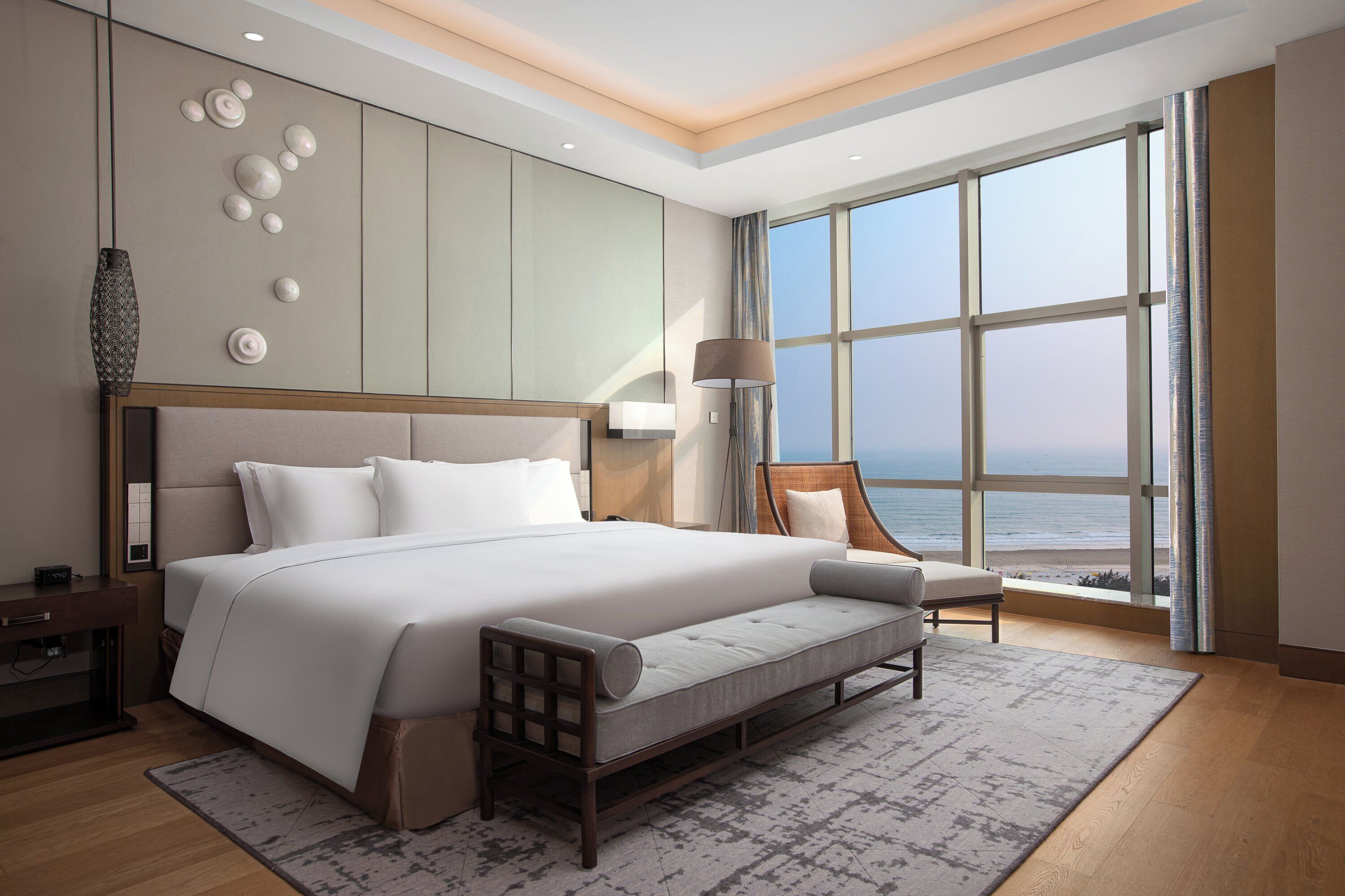 A modern hotel room with a large bed, a seating area, a view of the ocean, and a large window.