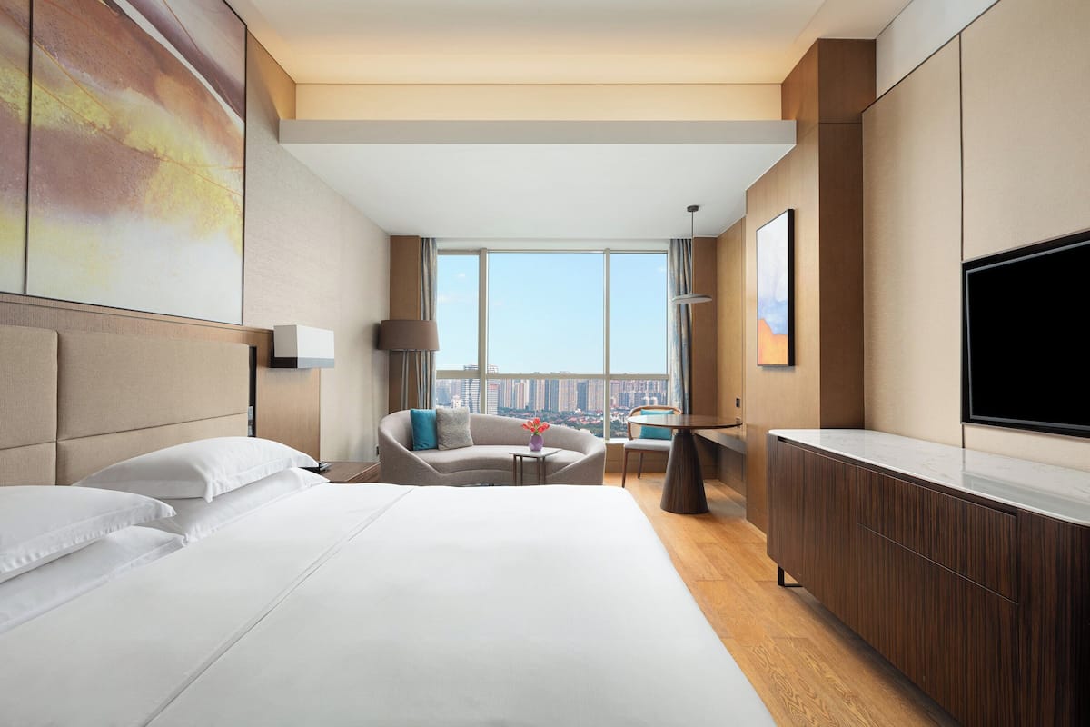 A modern hotel room with a large bed, a sofa, a small table, a TV, and a city view.