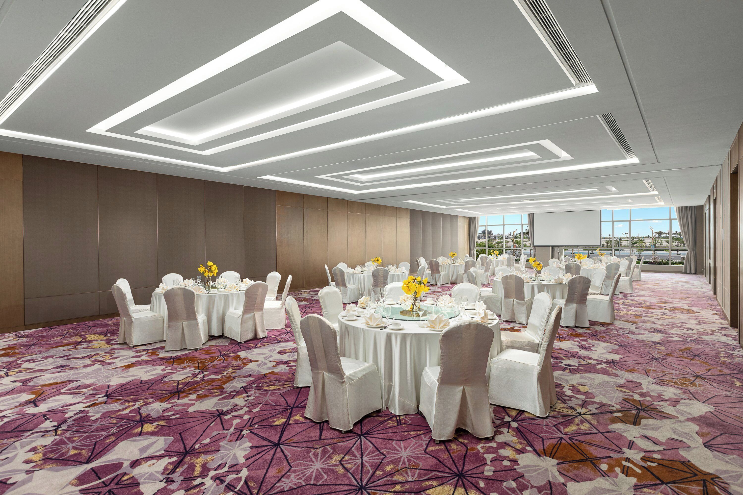 Un salón de banquetes con mesas redondas, sillas blancas y una alfombra estampada.