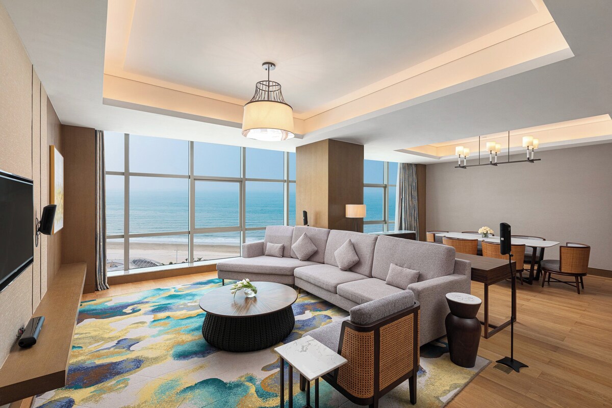 Habitación de hotel moderna con un ventanal grande que ofrece vista al océano, un rincón de descanso con sofá y sillones, una mesa de comedor y un televisor de pantalla plana montado en la pared.