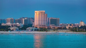 Un paisaje urbano costero con un hotel moderno destacado y una playa con gente y barcos.