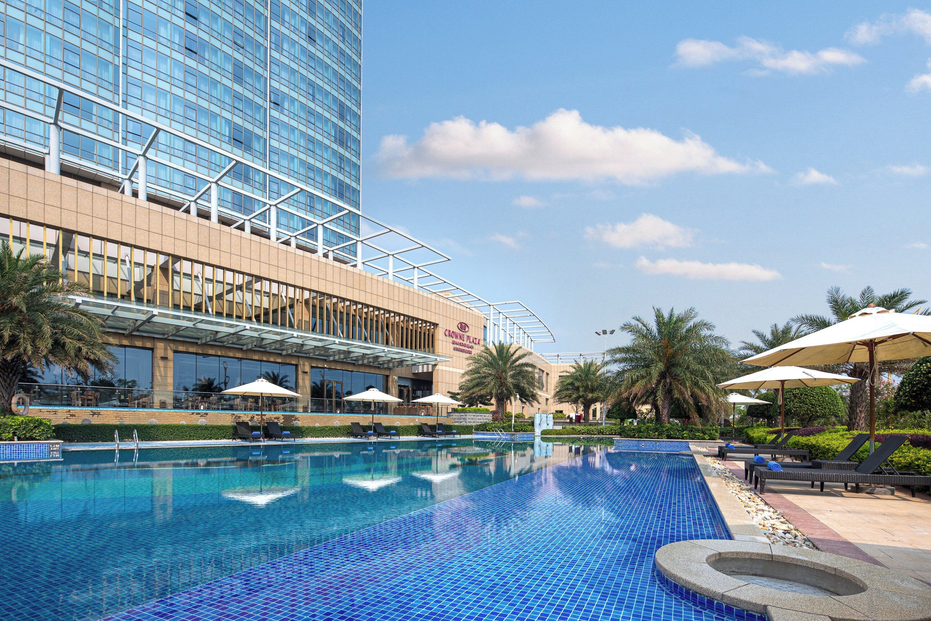 A hotel pool area with a large pool, lounge chairs, umbrellas, and a modern building in the background.