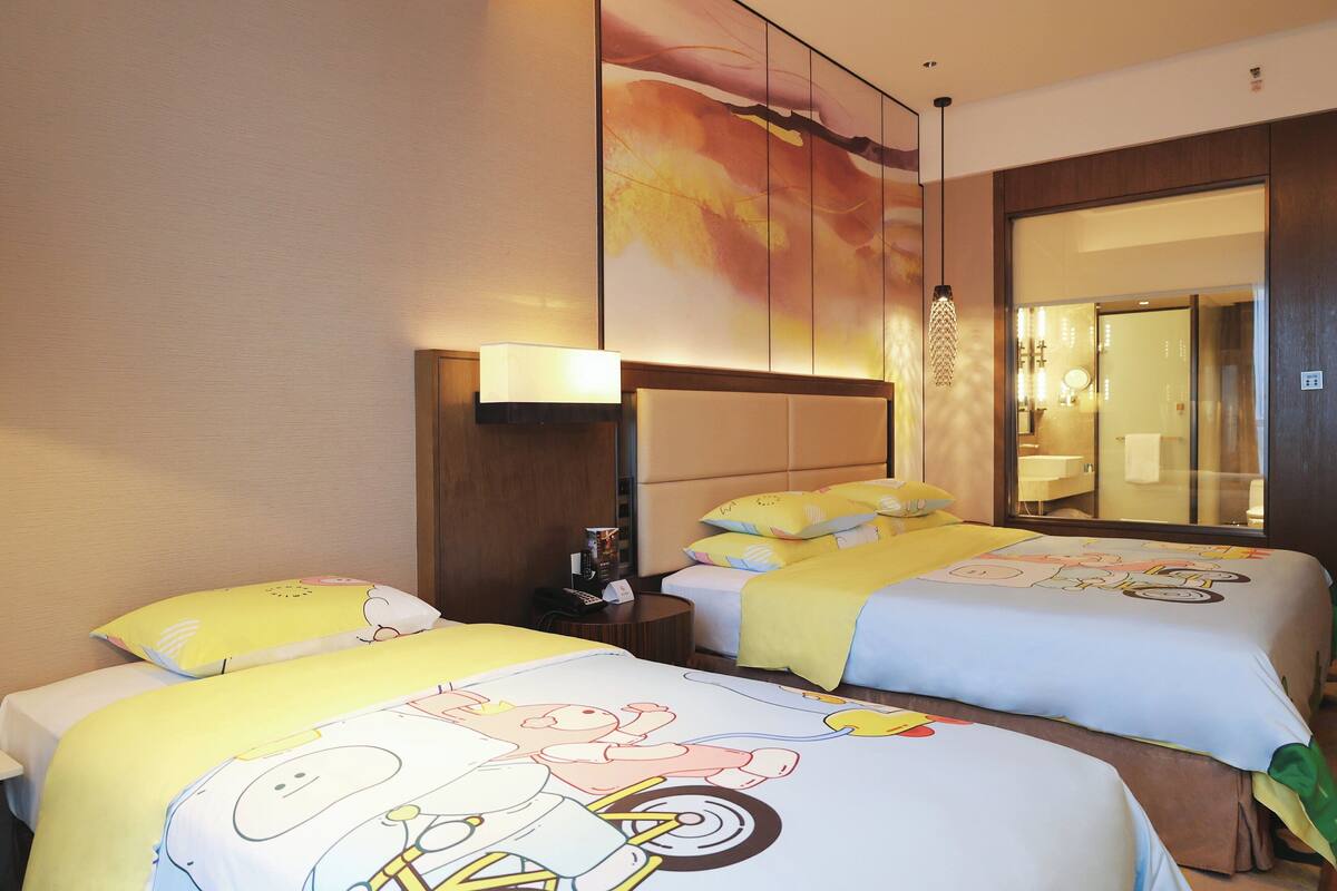 Habitación de hotel con dos camas, cada una con diseños de personajes de dibujos animados en la ropa de cama. Hay una mesita de noche con una lámpara, un espejo y una pequeña mesa con un teléfono y una revista. La habitación tiene un ventanal con vistas al exterior.