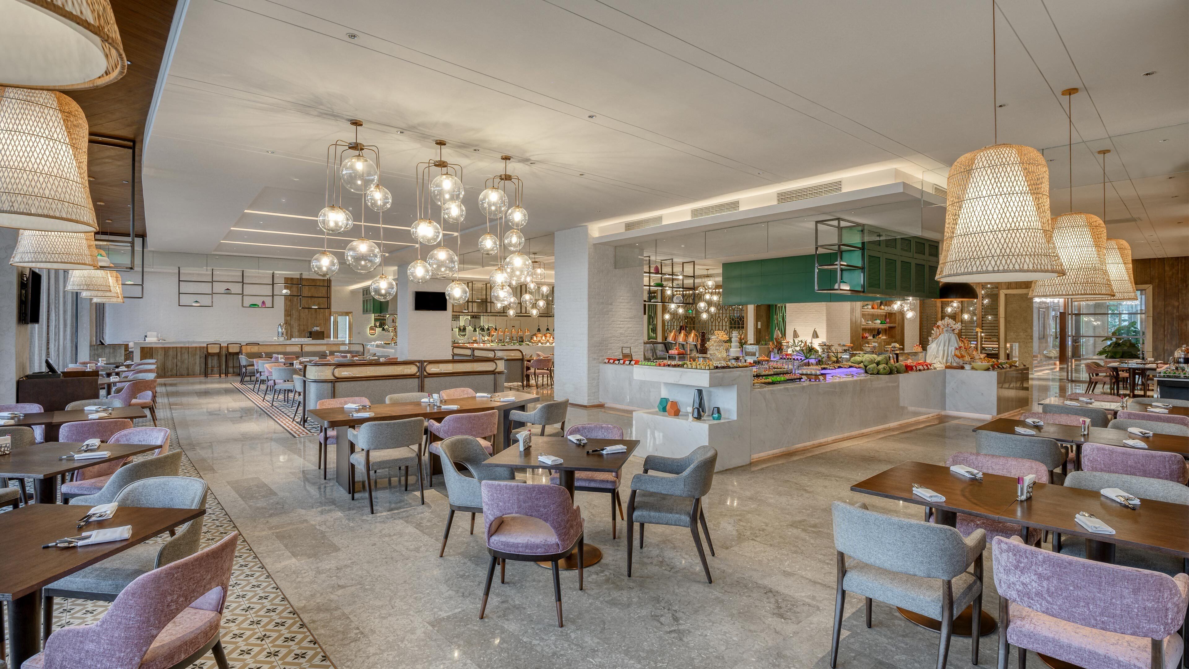 Un amplio vestíbulo de hotel con varias zonas de restauración, un bar central y un techo bien iluminado con lámparas esféricas colgantes.