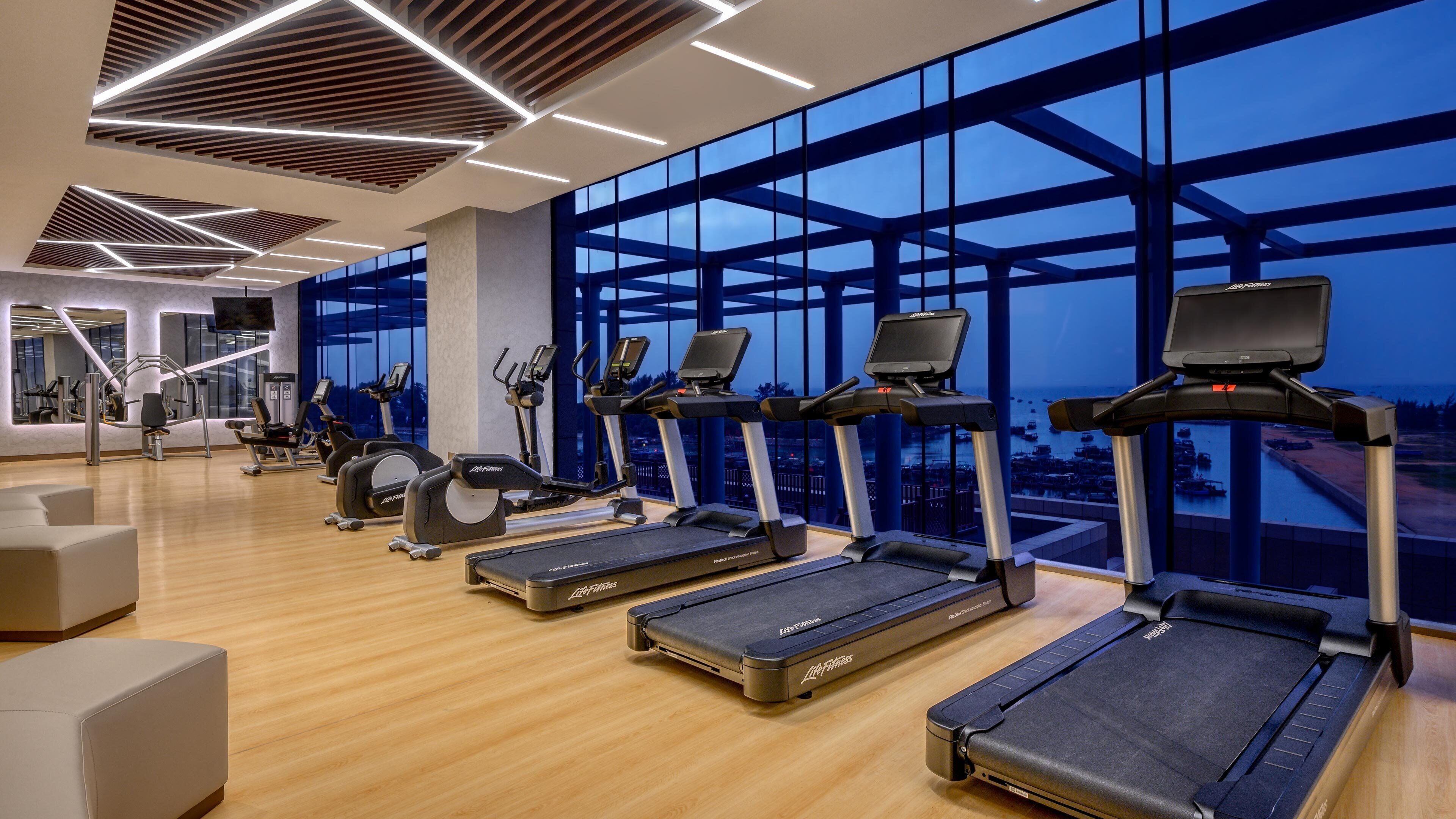 Un gimnasio moderno con cintas de correr, equipamiento de ejercicio y amplias ventanas con vistas a la ciudad.