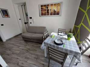 A dining area with a table set for two, a sofa, and a wall-mounted picture.