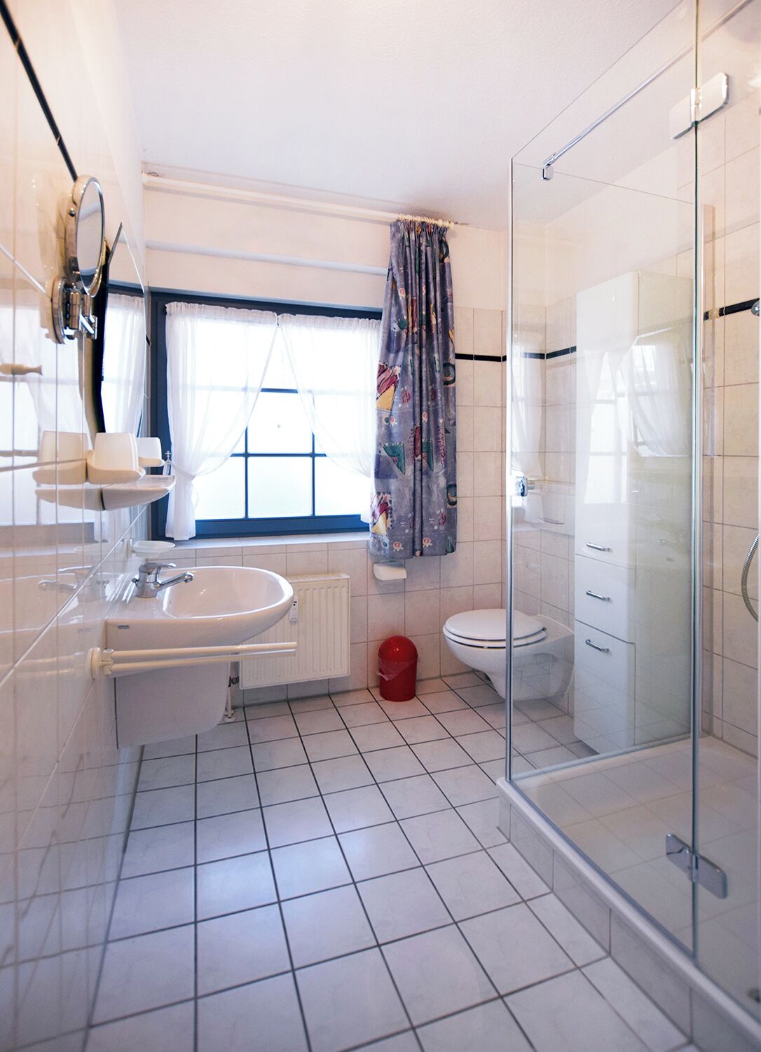 Bany modern amb plat de dutxa de vidre, un lavabo blanc, vàter i finestra amb cortina.