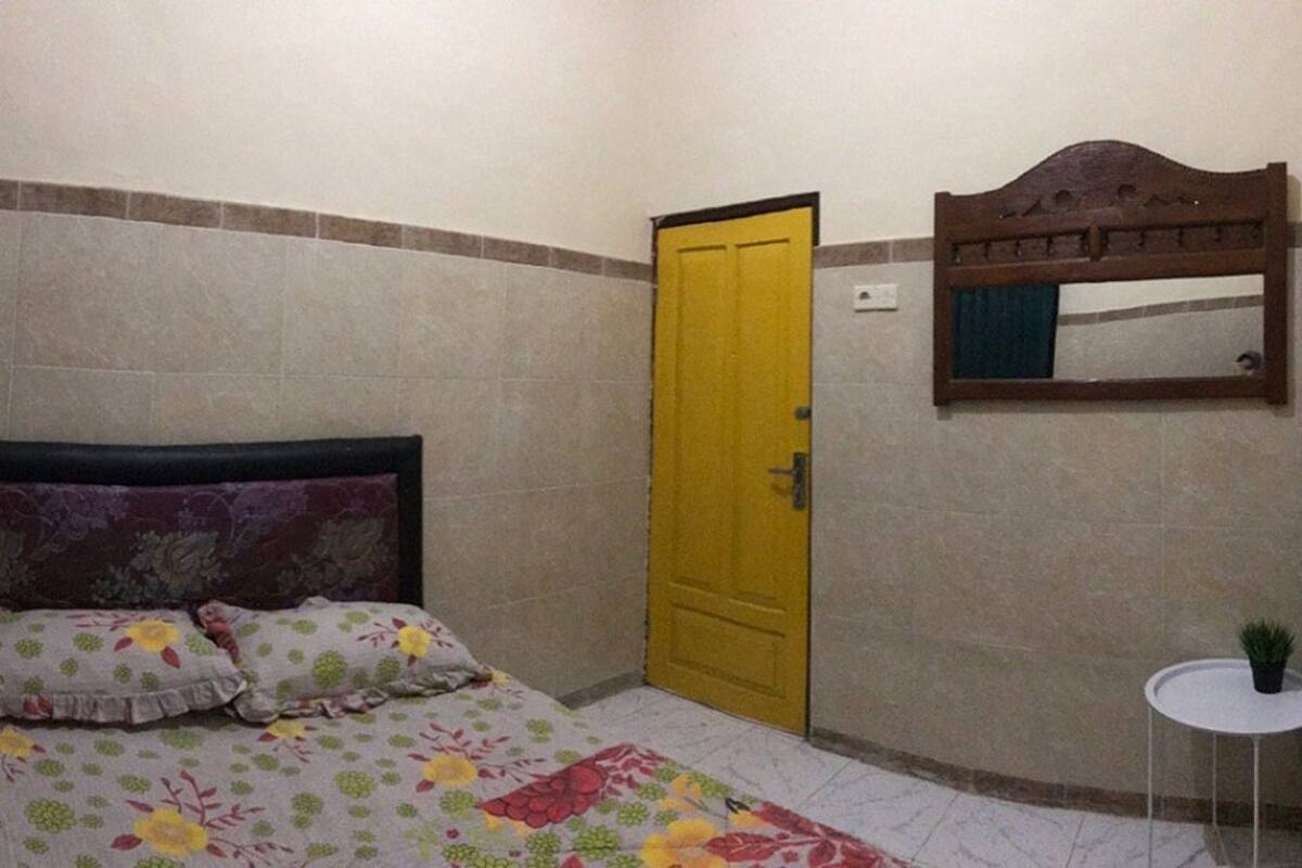 A small, neatly arranged bedroom with a bed, a yellow door, a small table with a plant, and a mirror on the wall.