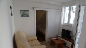 Une petite pièce bien éclairée, avec un canapé beige, un téléviseur, une petite table et une porte donnant sur une chambre à coucher.