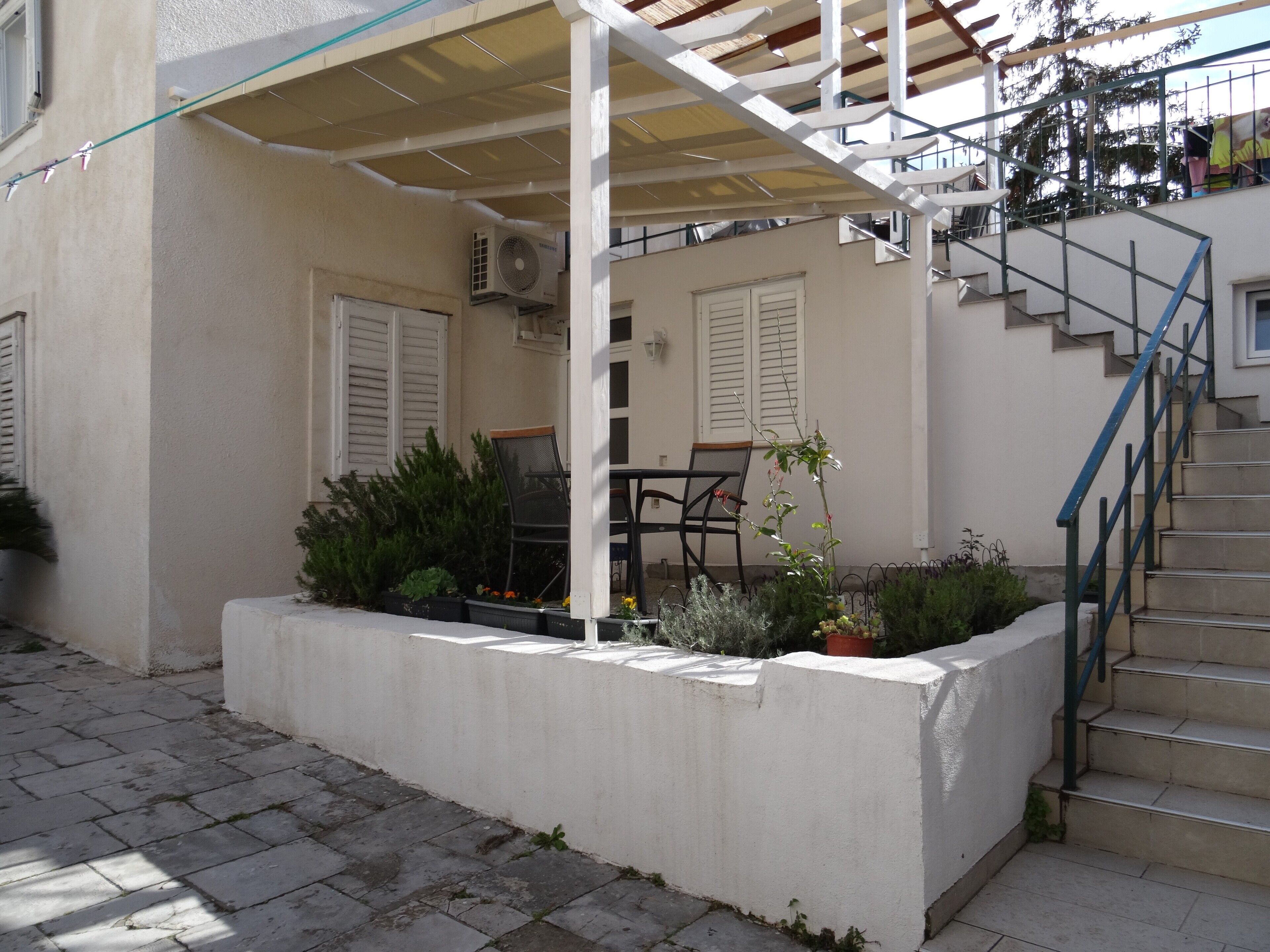 Un espace extérieur aménagé, abrité, avec une table et des chaises, un bac à plantes en béton agrémenté de végétaux, et un escalier menant à un niveau supérieur.