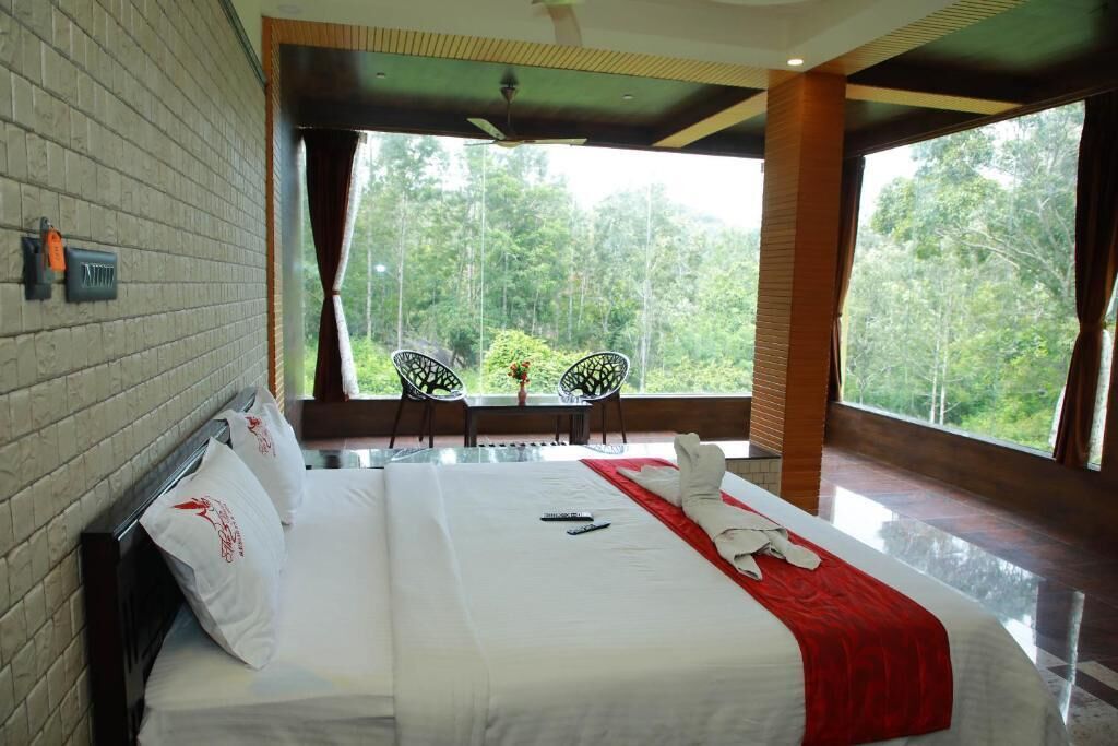A hotel room with a large bed, a view of the forest through the window, and a small table with chairs outside.