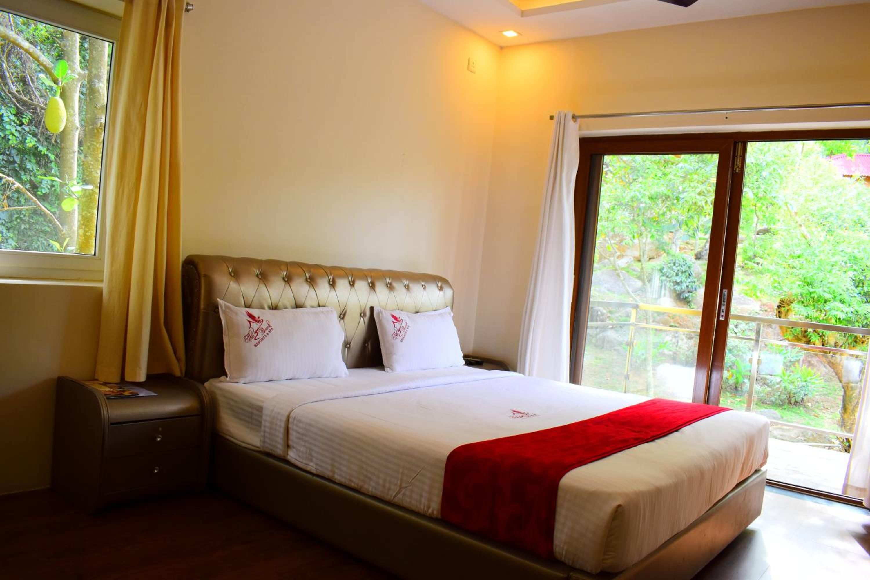 A hotel room with a bed, a nightstand, a window with curtains, and a view of greenery outside.