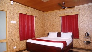 A hotel room with a wooden ceiling, a bed with a red and white bedspread, red curtains, a ceiling fan, and a coffee maker on a shelf.