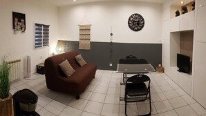 A compact living space with a brown sofa, a glass dining table, a black TV, and a wall clock. - Studio LE COLONEL, in the heart💖of the beautiful village of Marseillan  (Marseillan)