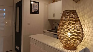 A modern kitchen with white cabinets, a white countertop, a hanging decorative heart, and a textured wall lamp. - Studio LE COLONEL, in the heart💖of the beautiful village of Marseillan  (Marseillan)