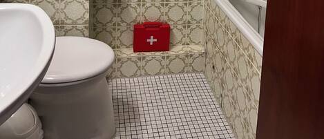 Et badeværelse med mønstret fliser, et toilet, en bruser og et rødt førstehjælpskit.