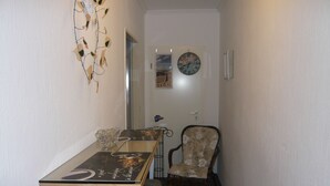 Un pasillo estrecho con una mesa, dos sillas, un adorno de pared, un reloj y una puerta que da a otra habitación.