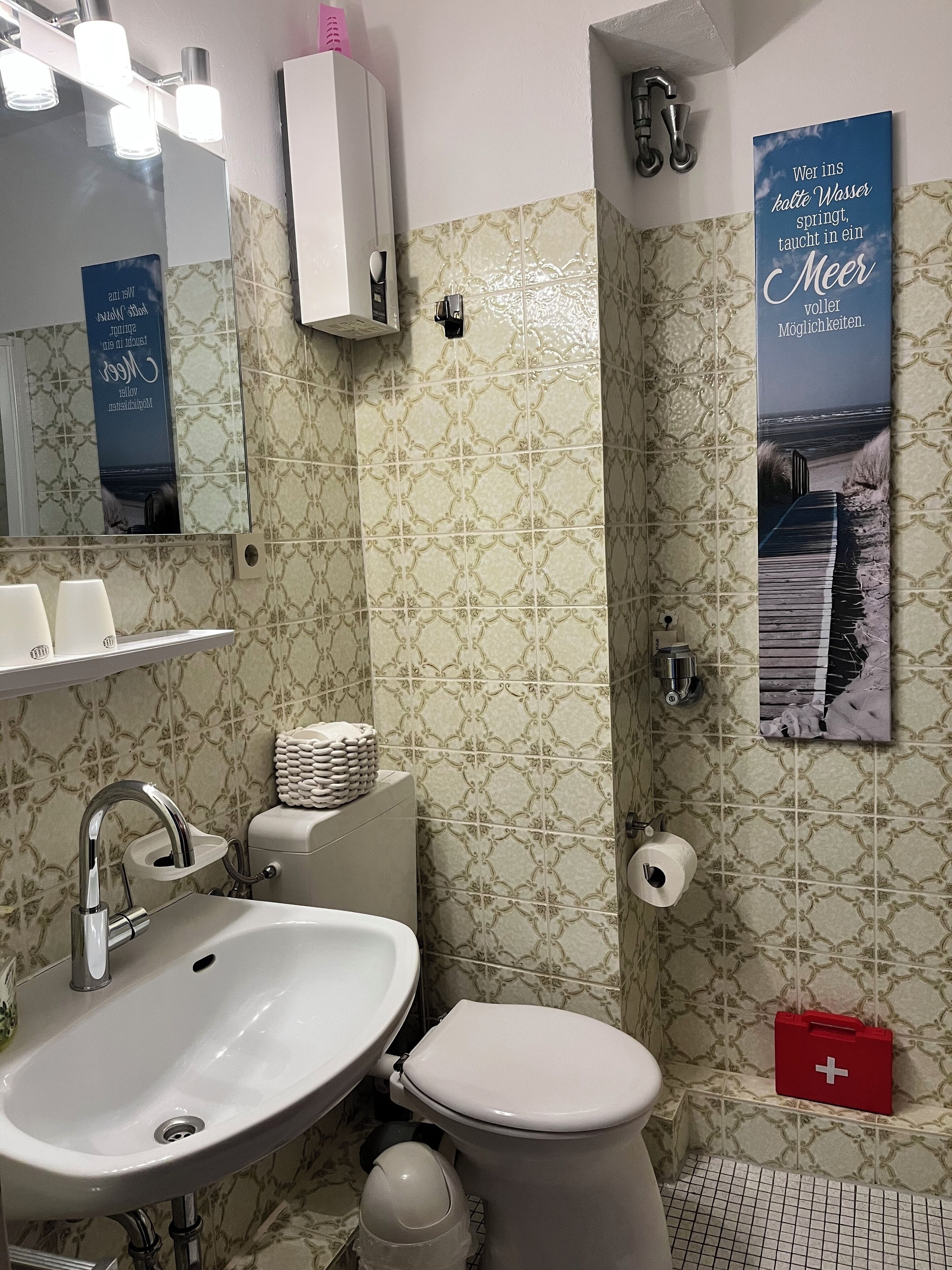 Une salle de bain avec un lavabo, des toilettes et une douche. On y trouve un miroir, un porte-serviettes et une affiche accrochée au mur.