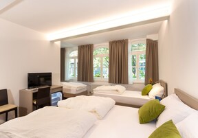 A hotel room with two beds, a desk with a computer, and large windows with curtains.