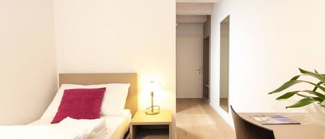 A hotel room with a bed, bedside table with a lamp, a mirror, a desk, and a plant.