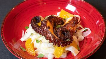 A red bowl containing grilled octopus, orange segments, and white noodles.