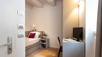 A compact hotel room with a bed, bedside table, chair, desk, and a TV.