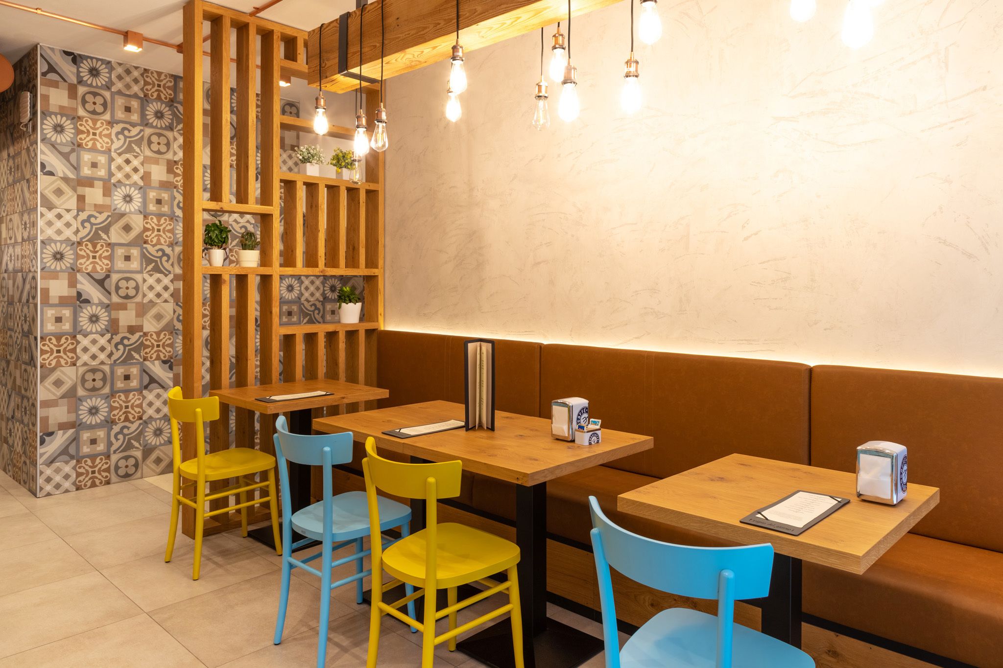 Interior de restaurante com mesas e cadeiras de madeira, parede estampada e lâmpadas penduradas.