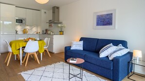 A modern living room with a blue sofa, a dining table with chairs, a kitchen area, and a framed artwork on the wall. - Large studio, feet in the water! (Bénodet)