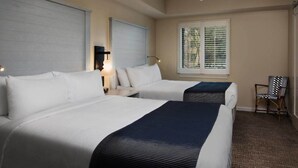 Ein Hotelzimmer mit zwei Betten, einem Deckenventilator, einem Fenster mit Jalousien, einem Sessel und einem Nachttisch mit Lampe.
