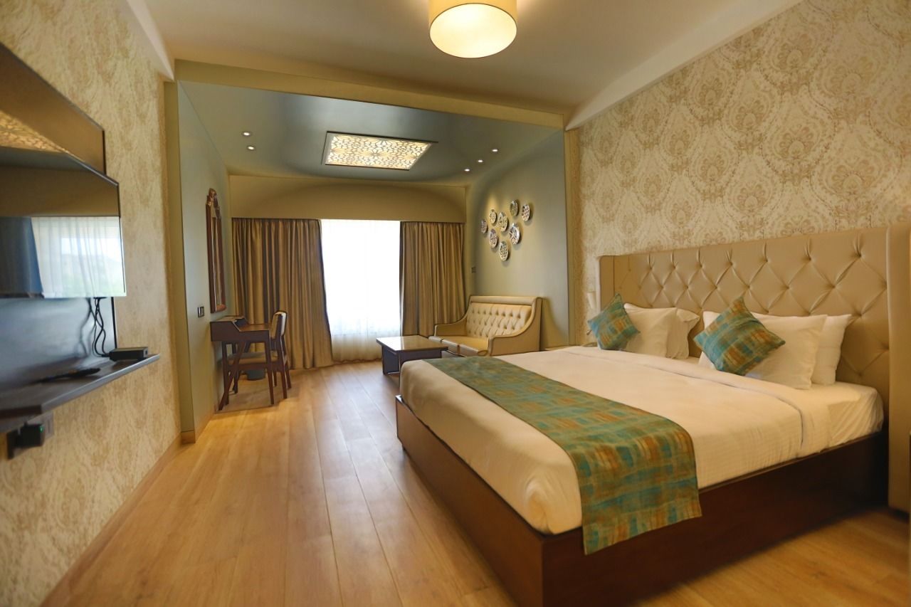A hotel room with a large bed, a desk, and a chair. There is a window with curtains, a ceiling light, and a wall-mounted TV.