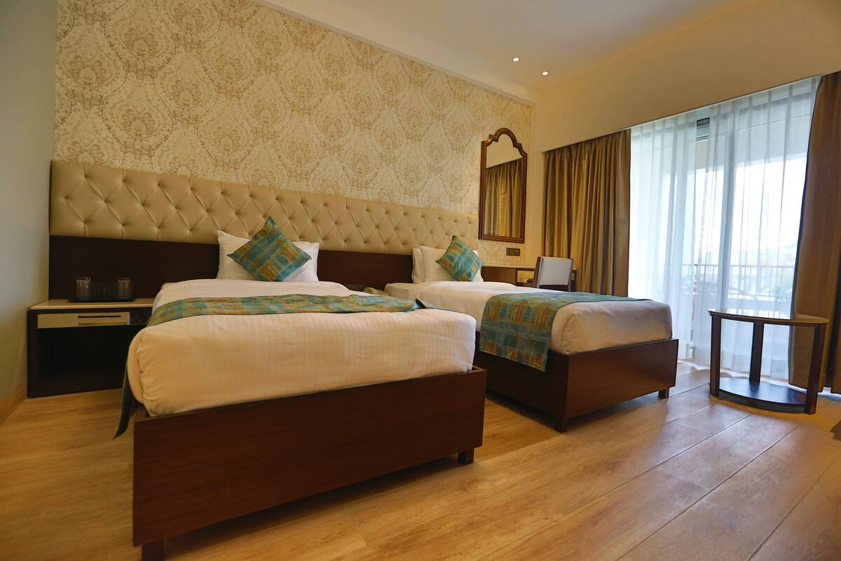 A hotel room with two beds, a large mirror, a bedside table, and a view of the outdoors through sheer curtains.